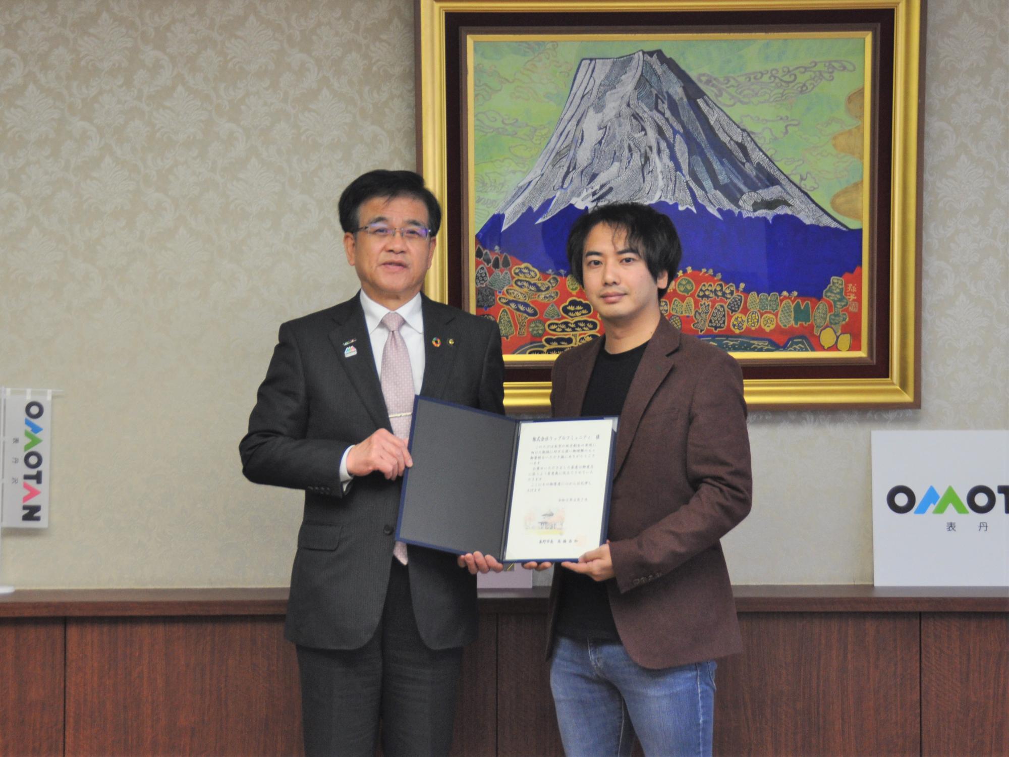 富士山の絵画を背景にした室内のカウンター前で、市長と株式会社リップルコミュニティの関係者が、中央で証書のような書類を広げて持ち、カメラに向かって真剣な表情で立っている記念写真
