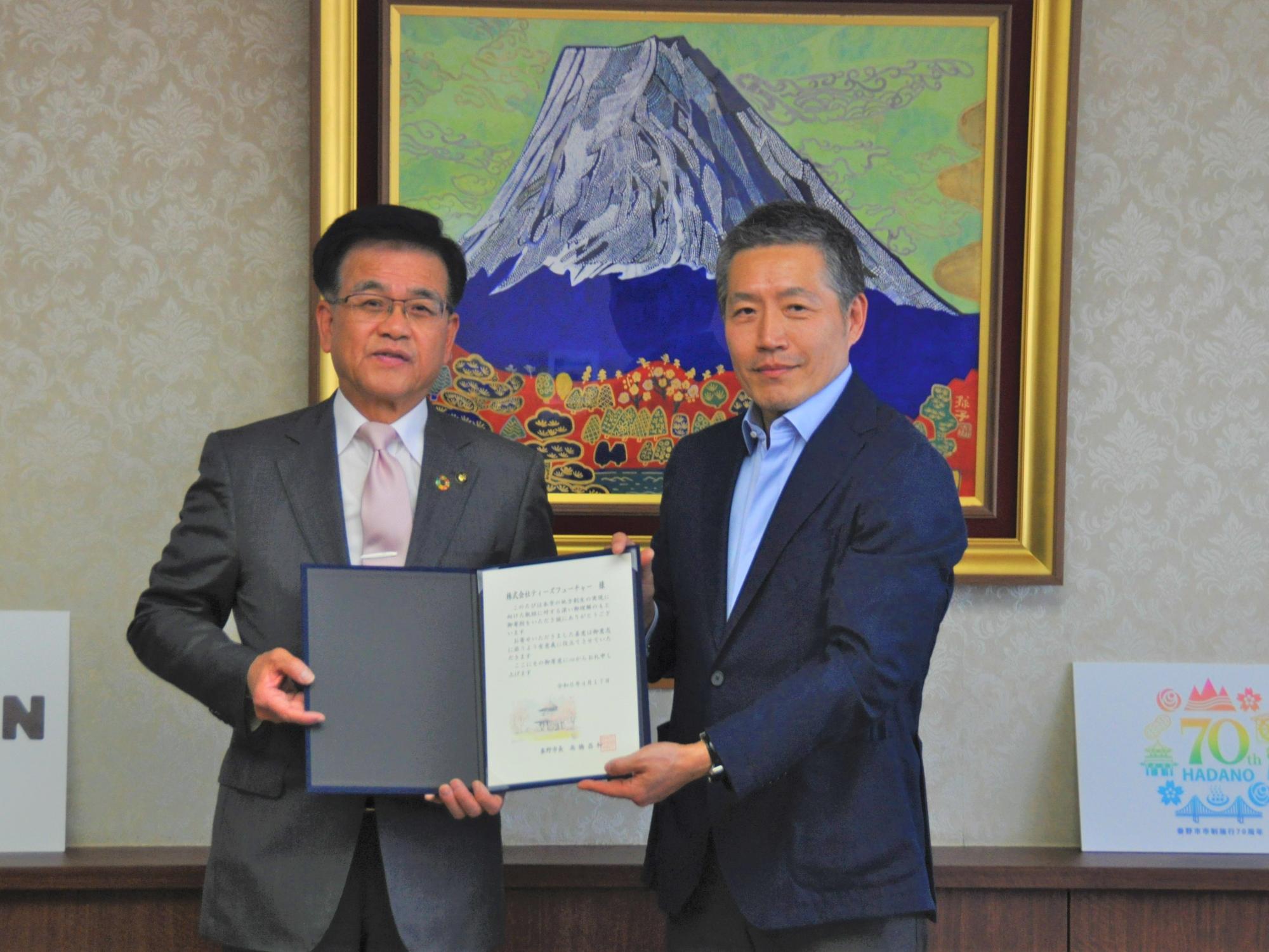 金色で縁取られた富士山の絵画が背景の室内のカウンター前で、ダークスーツとシャツ姿の男性と市長が、中央で開いた賞状のような書類を丁寧に持ってカメラに向かって立っている記念写真