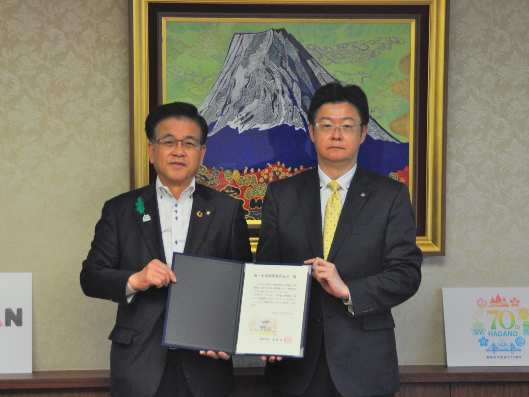 富士山の大きな絵画が飾られた室内の木製カウンター前で、スーツ姿の2人の男性が、中央で証書のような書類を広げてカメラ目線で立ち、左の市長は胸に緑色のバッジを付けている記念写真