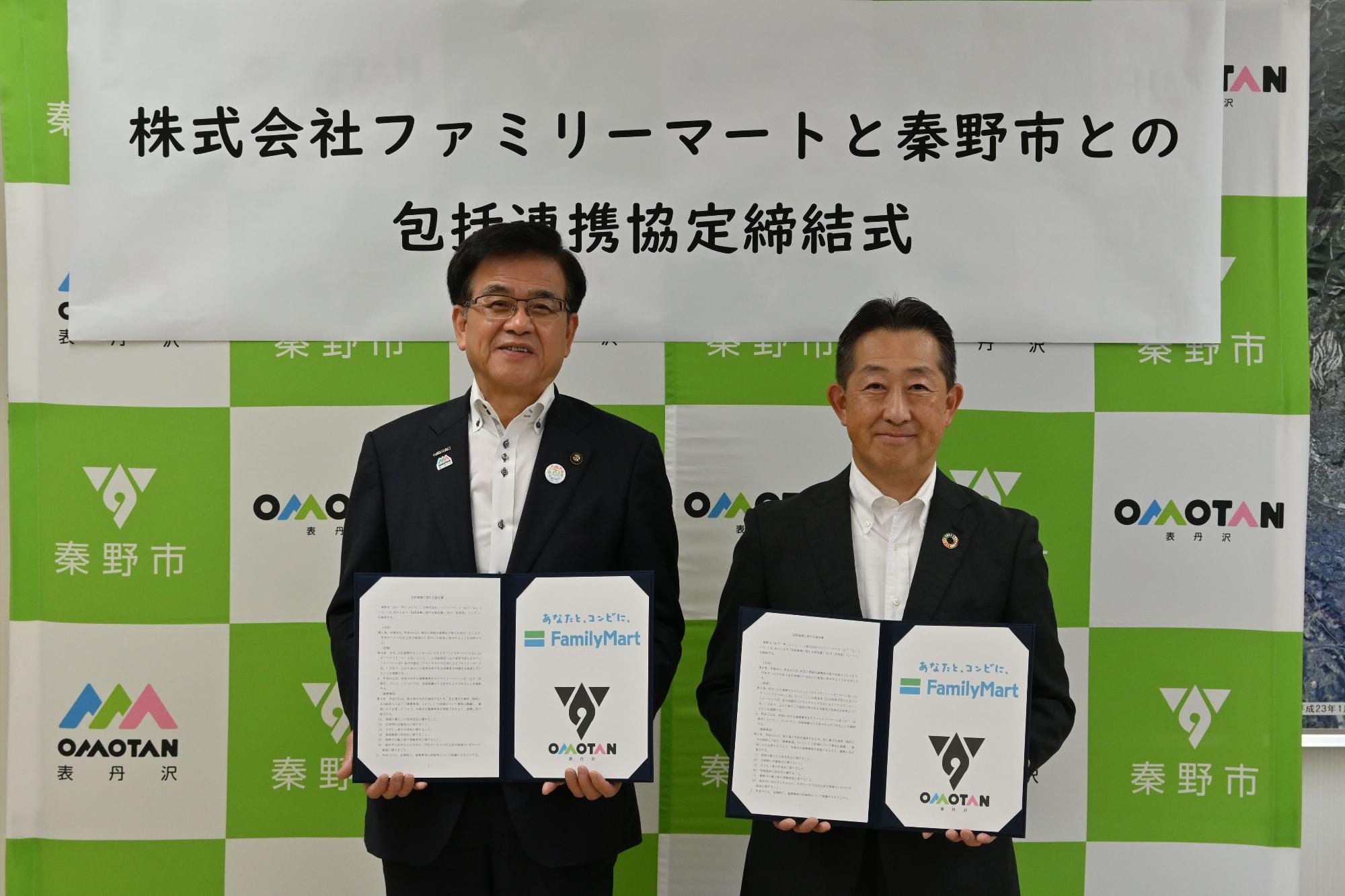 秦野市のバックボードの前で高橋市長とファミリーマートの関係者の男性が協定書を持ち記念撮影をしている写真