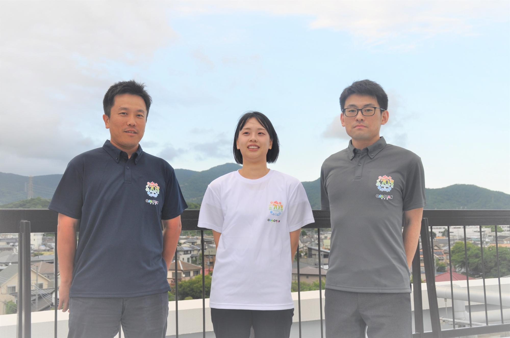 手すりの前で、オリジナルのカラフルなロゴが入ったポロシャツやTシャツを着用した男女三人が、曇り空の下で街並みと奥に連なる山々を背景に、真正面を向いて立っている集合写真