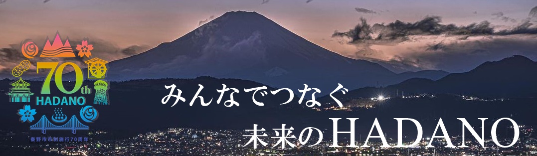 秦野市の街並みの向こうにそびえ立つ富士山のシルエットを背景に、「みんなでつなぐ 未来のHADANO」という白い文字と、「秦野市市制施行70周年」を記念するロゴマークが左側に配置された画像