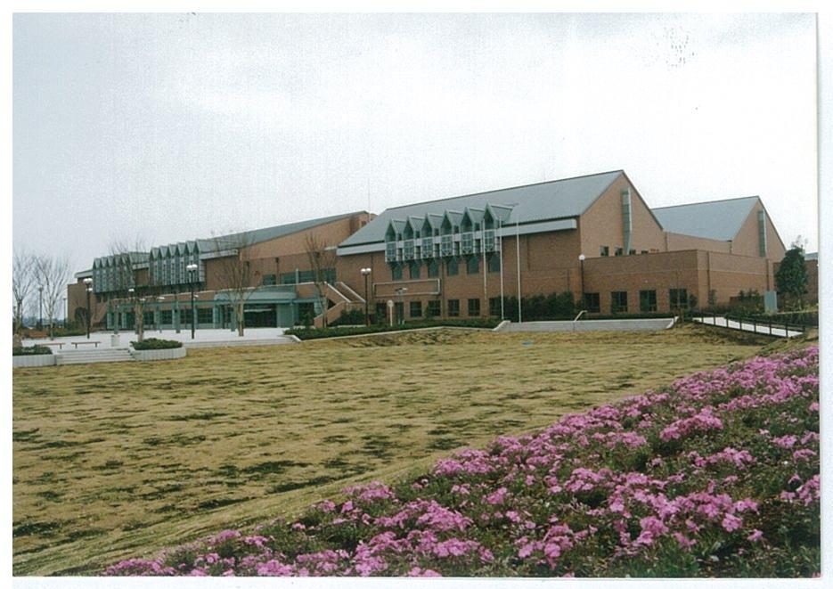 なだらかな芝生の広場とピンク色の花が手前に広がり、奥にはレンガ造りの重厚な外観で、多くの屋根と窓が特徴的な総合体育館を捉えた遠景写真