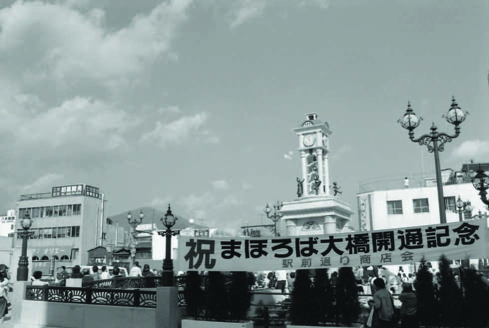 大勢の人が集まる広場の中央に、「祝 まほろば大橋開通記念 駅前通り商店会」と書かれた横断幕が掲げられている、開通記念式典を捉えた白黒写真