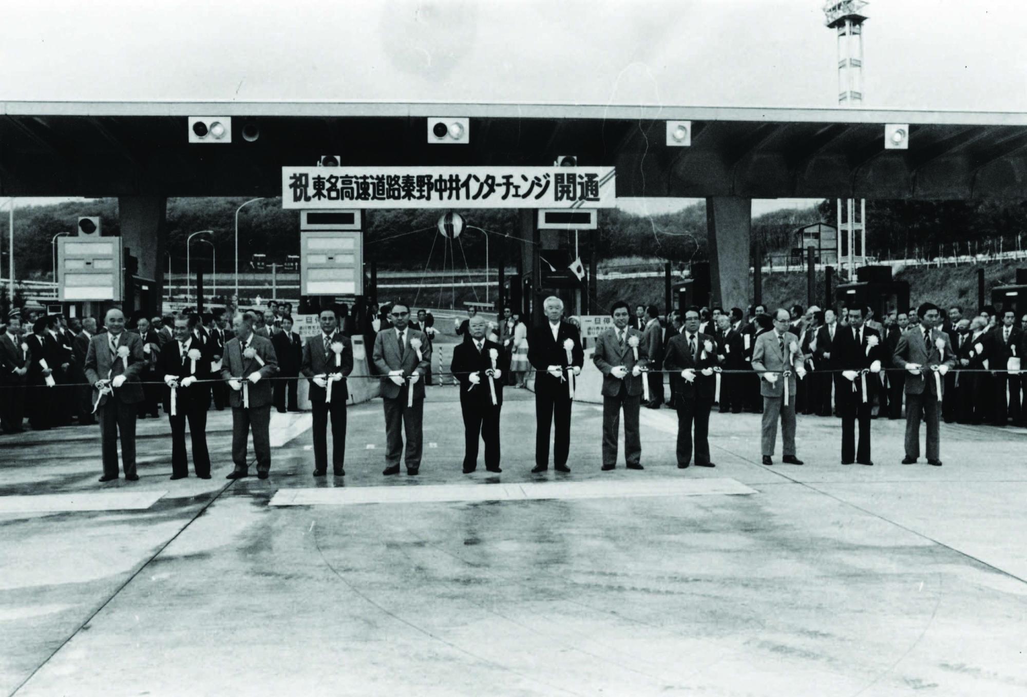 「祝 東名高速道路秦野中井インターチェンジ開通」の横断幕の下で、多くの関係者が並び、中央で数人がテープカットを行なっている開通記念の白黒写真