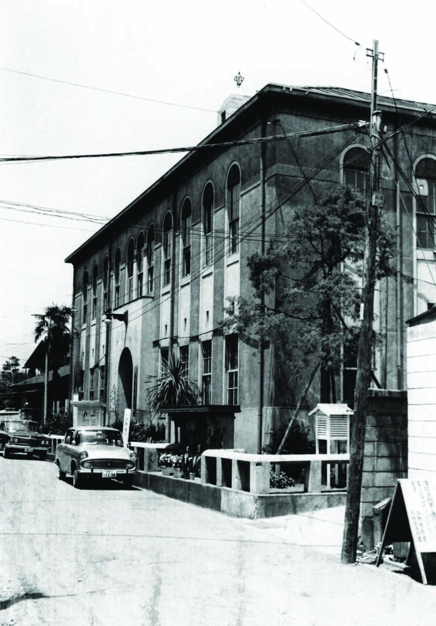 昭和時代の街並みを背景に、縦長の丸窓が特徴的なルネサンス様式を思わせる洋風の建物が建ち並ぶ道路の様子を、電柱と車も写り込んだ状態で捉えた白黒写真