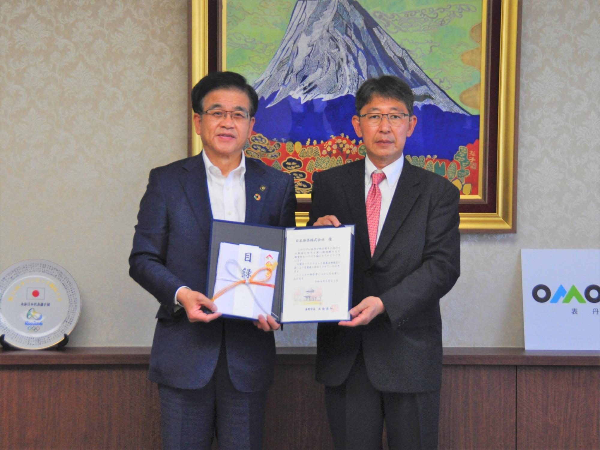 富士山の絵画を背景にした室内のカウンター前で、市長と株式会社トープラ様が、中央で証書のような書類を広げて持ち、カメラに向かって真剣な表情で立っている記念写真