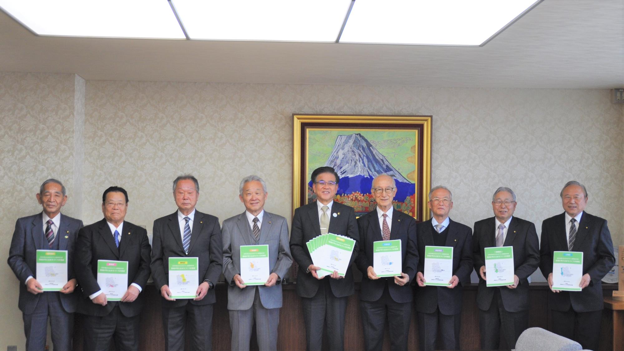 応接室のような部屋で、スーツ姿の9人の男性が、緑色の表紙の冊子を手に持ち一列に並んでいる集合写真