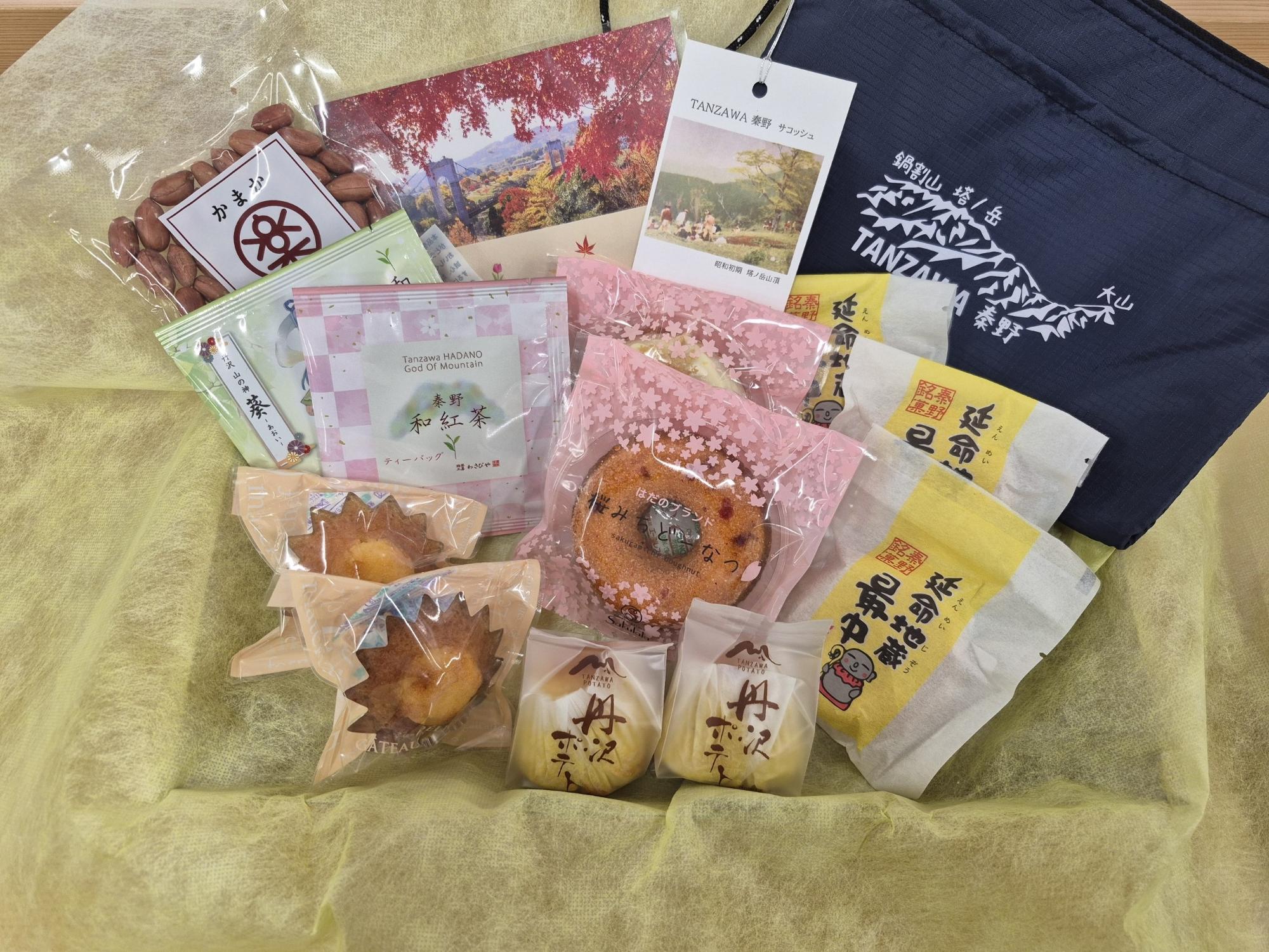 淡い黄色のラッピングペーパーの上に、濃紺のロゴ入りバッグと紅葉のポストカードやパンフレット、そして秦野名産の落花生や「鶴寿」と書かれた黄色のパッケージの「丹沢お豆っこ」、ドーナツやマドレーヌなどの様々な焼き菓子が多数並べられた、地域の特産品やお土産品の詰め合わせギフトの写真