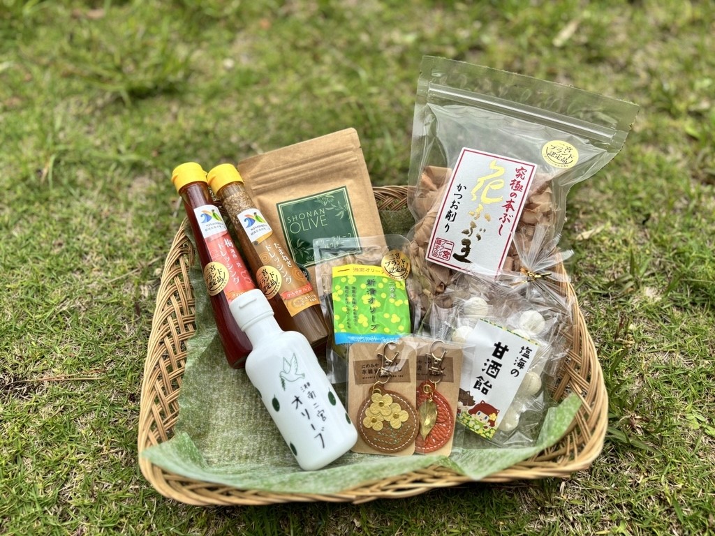緑の芝生の上に置かれた籠の中に、湘南オリーブオイル、レモン風味のドレッシング、柑橘系の調味料、柑橘類のキーホルダー、甘酒の飴、二宮産かつお節「花の舞」など、地域の特産品や調味料が詰め合わせられたギフトセットの写真