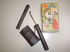 焦げ茶色の革製と思われる煙草入れと、同じく焦げ茶色の筒状の煙管筒と煙管、そして右側には浮世絵のような絵柄のマッチ箱が置かれている写真