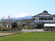 雪をかぶった富士山を背後に、水田や木々のあるのどかな風景の中、白壁に黒い木組みの伝統的な造りの小さな水車小屋と2階建ての建物が並んで立っている、ふるさと伝承館の外観写真