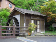 道路沿いの欄干の後ろに、木々に囲まれた木造の古い水車小屋が立っている様子の写真