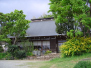 伝統的な造りの平屋の建物の手前左右に、大きな木や低木の植栽がある様子の写真