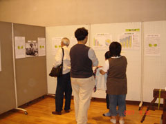 展示会場で、4人の来場者が壁に掲示されたグラフや文章のパネルを熱心に見ている様子の写真