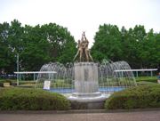 広場の中央にある噴水に、上半身裸の人物と動物がモチーフになった像が設置され、周囲を木々と低木が囲む中央運動公園の屋外写真