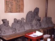 木の台の上に、複数の古い石像が並んで安置され、後ろの壁に縦書きに文字が書かれた茶色の紙が貼られている石造十王像の写真