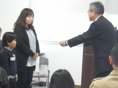 スーツ姿の男性と向かい合わせに黒いカーディガンを羽織った女性と、その隣にトレーナーを着た男の子の親子に、表彰状渡している表彰式の写真