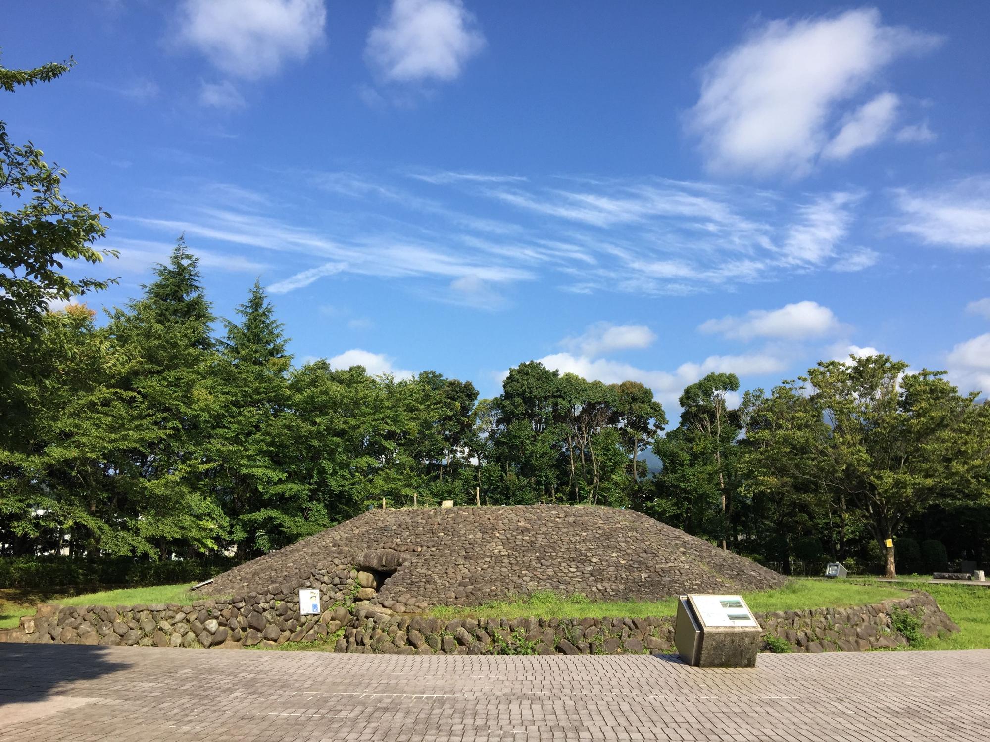 緑の木々を背景に、周りを低い石垣で丸く囲まれた、石を積み上げた円墳があり、円墳の入口が石垣につながっている様子がわかる復原古墳（1号墳）の写真