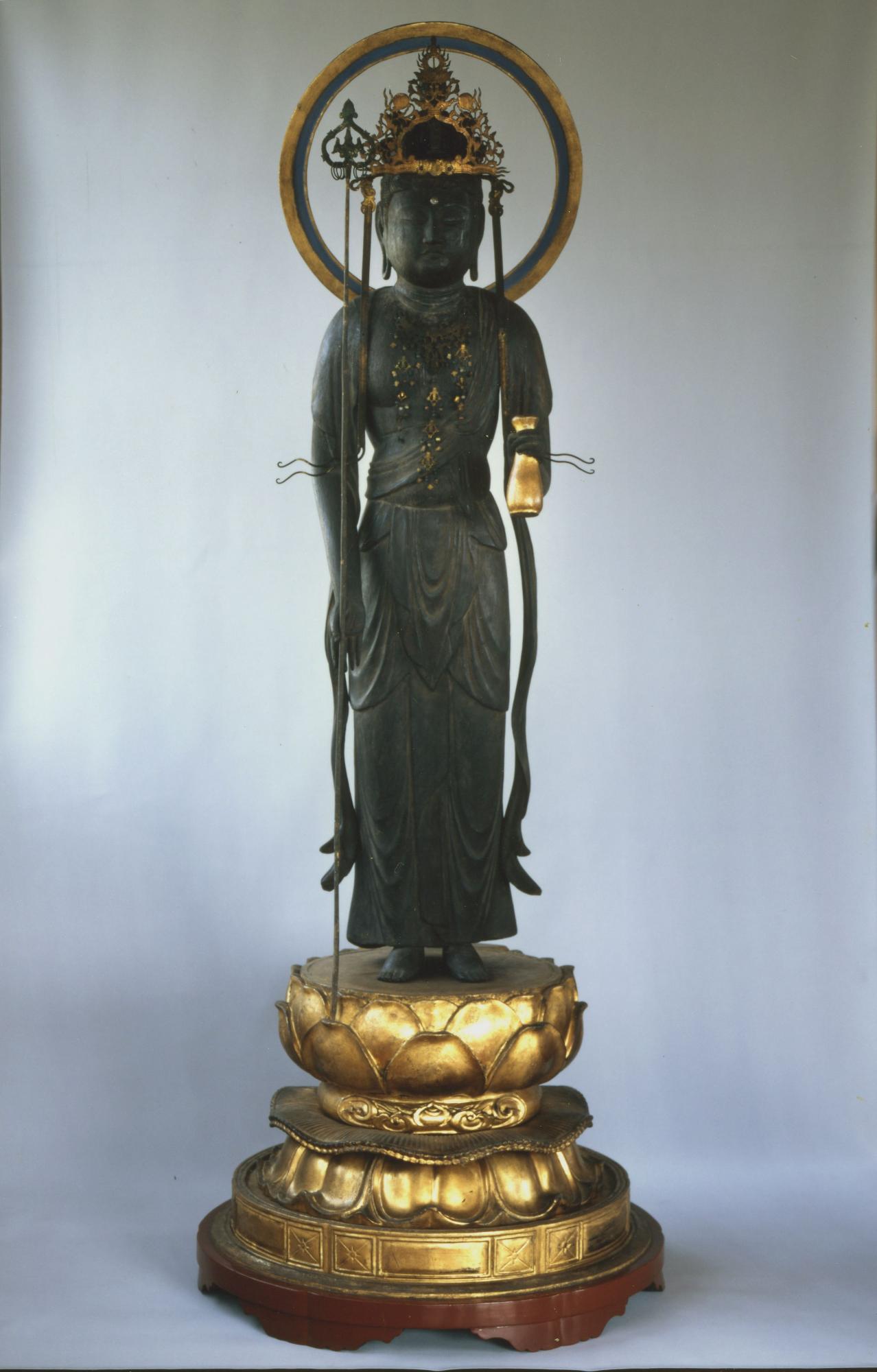 豪華な宝冠と円光を背負い、金色の蓮華座の上に立つ、左腕が欠損し銅色の装飾が施された古い仏像が、正面から撮影された写真