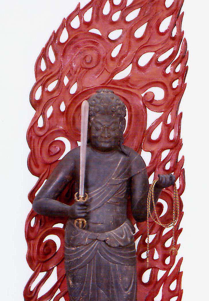 激しい炎の形をした赤い光背を背負い、右手に剣、左手に羂索を持つ、険しい表情の仏像を正面から捉えた写真