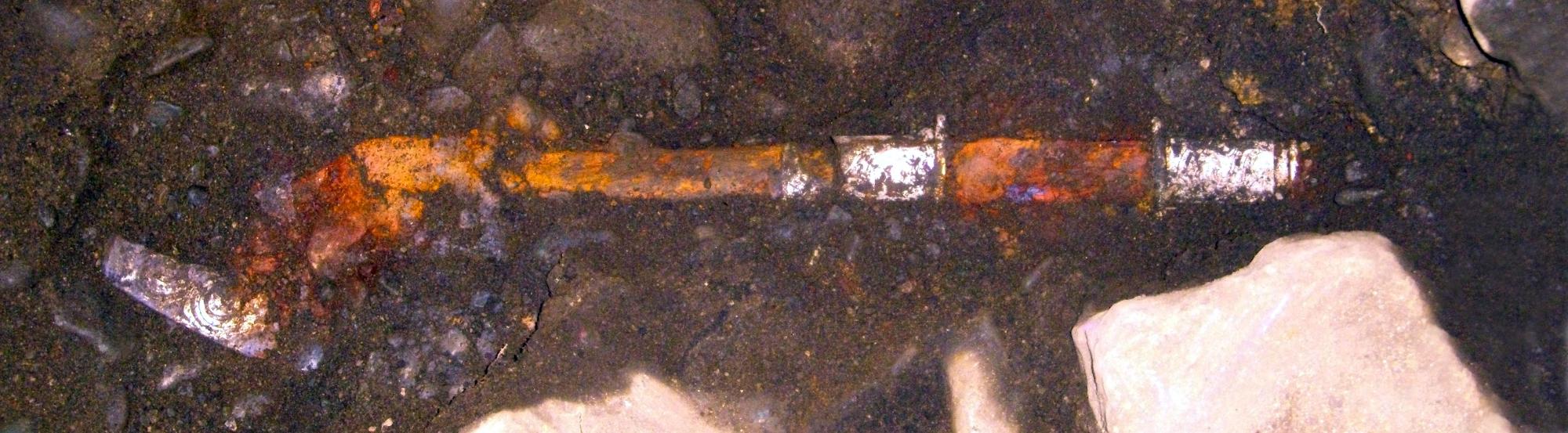 暗く湿った土の中に横たわる、錆びて茶色や白に変色した金属製の古い刀の一部を接写した写真