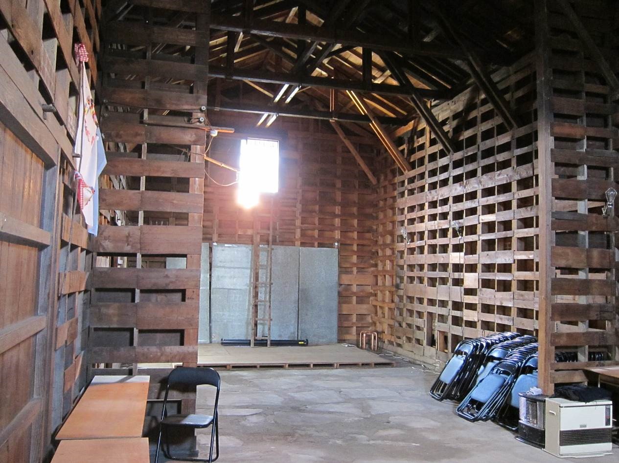 パイプ椅子が積み重ねられ、奥にはすのこが設置されている剥き出しの木の柱や天井の梁が見える広い倉庫内部の写真