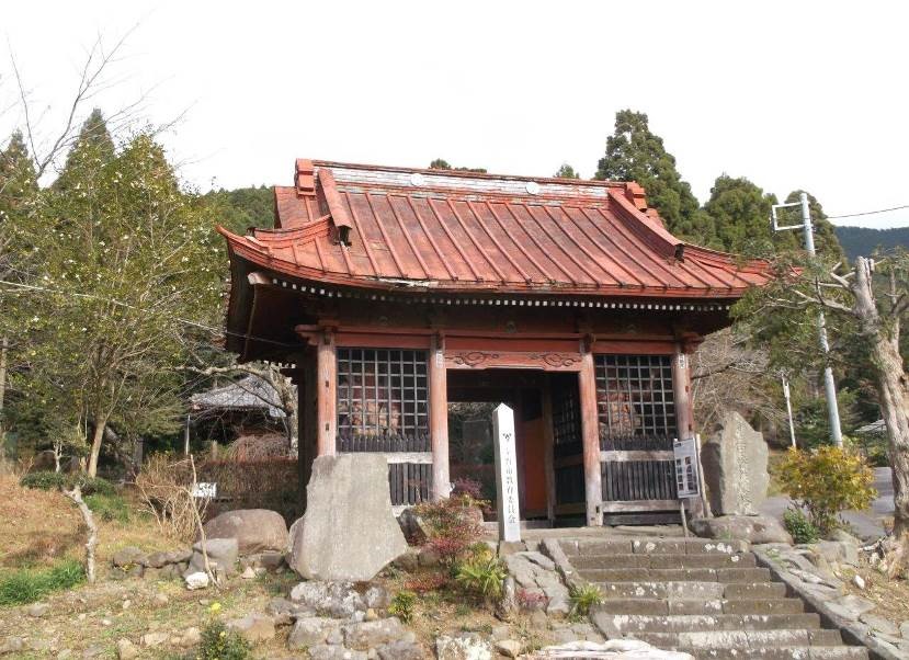 石段を登った先にある、手前に複数の石碑が立てられた、朱色の屋根と木製の格子戸が特徴的な仁王門の外観写真