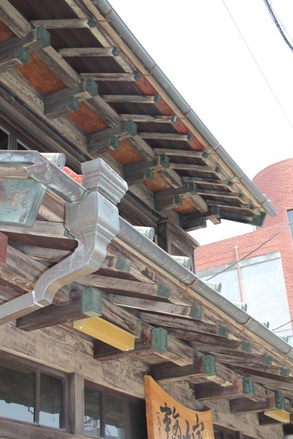 古びた木材が組み合わされた構造と、金属製の重厚な雨樋を軒下を下からクローズアップで写した写真