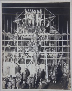 木造の足場を組まれた建物の前に、大勢の作業員や関係者が集まり、上部には旗のような装飾が見える棟上げ式の白黒の集合写真