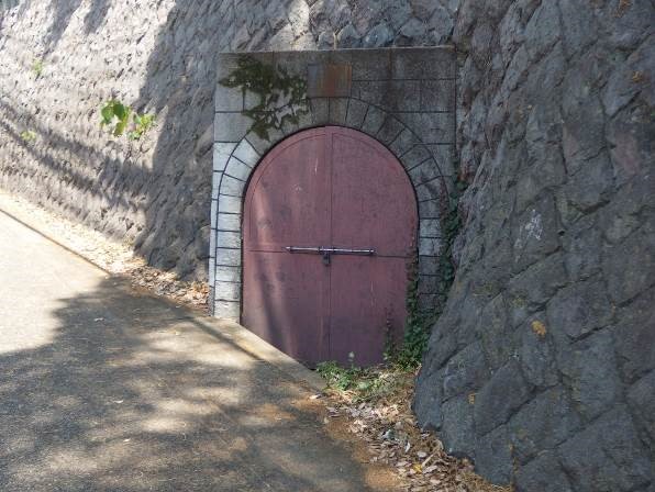 石垣に囲まれた斜面に埋め込まれて何かを収納する為の倉庫のようなアーチ状のレンガで縁取られた鉄扉を写した写真