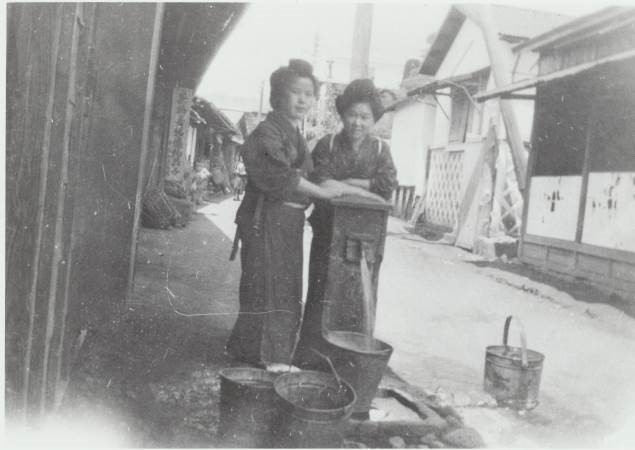 着物を着た二人の女性が、古い井戸ポンプの横に立ち、足元のバケツに水を入れている様子を写したモノクロ写真