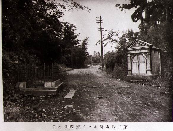 木々が生い茂った土砂で舗装されていない道の左側には墓のようなコンクリートの囲いが、右側には洋風の小さな小屋が建っている古いモノクロ写真