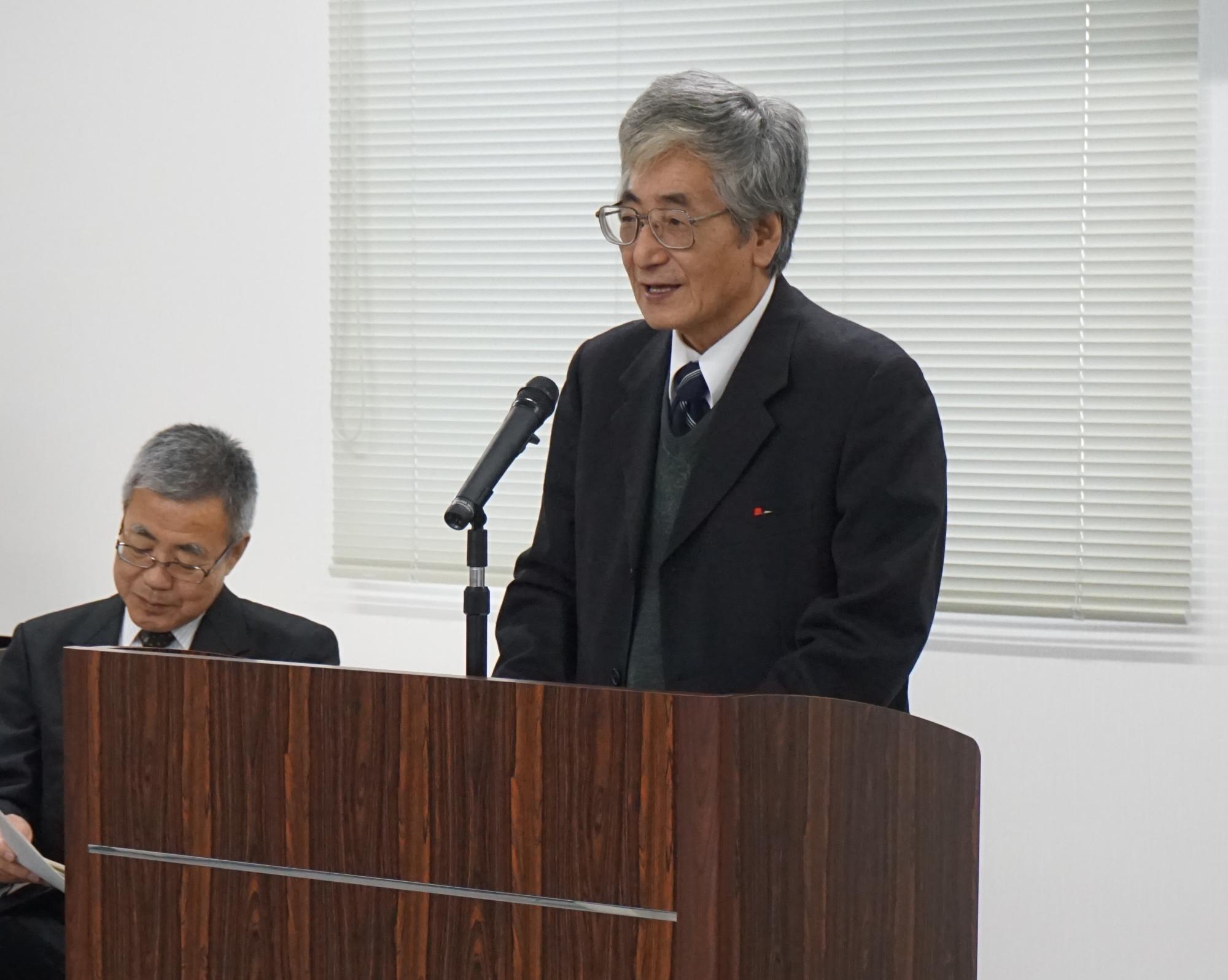 眼鏡をかけたスーツ姿の鍛冶実行委員長兼審査委員長が、濃い茶色の木目調の演台の後ろで話をしている写真