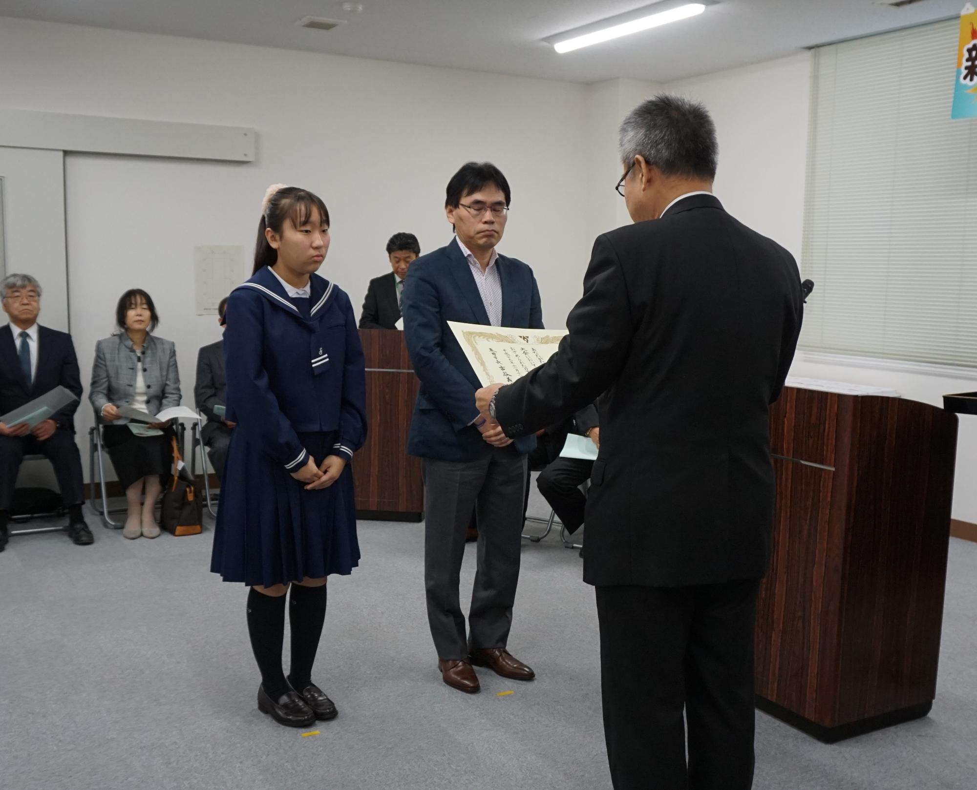 表彰式会場でセーラー服姿の女子学生とダークブルーのジャケットを着た男性の親子と向かい合わせにスーツ姿の男性が賞状を読んでいる写真