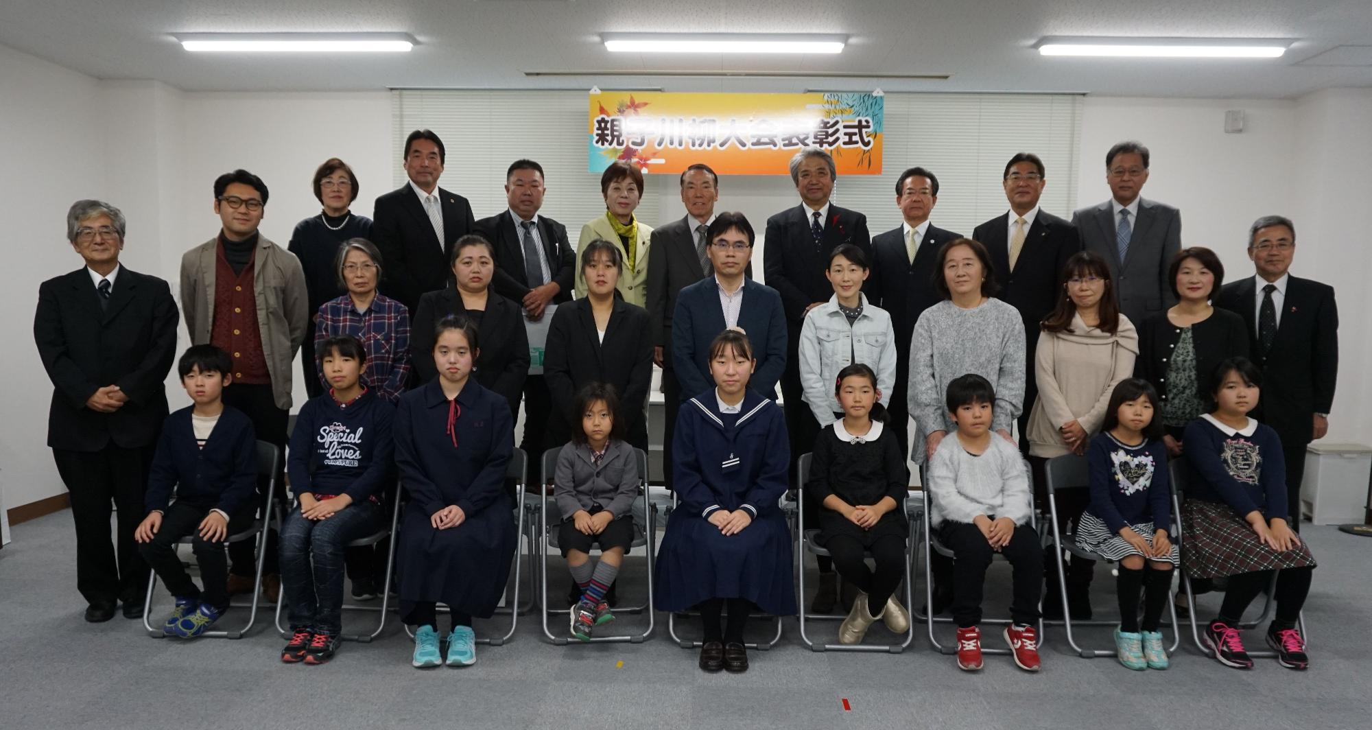 「親子川柳大会表彰式」の横断幕を背景に、保護者と関係者が後列に立ち、子供たちが前列の椅子に座っている集合写真