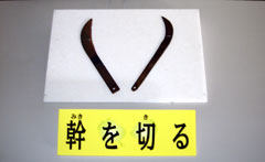 「幹を切る」と書かれた札が置かれ、左右対称の鎌のような刃物が2つ並べられている写真