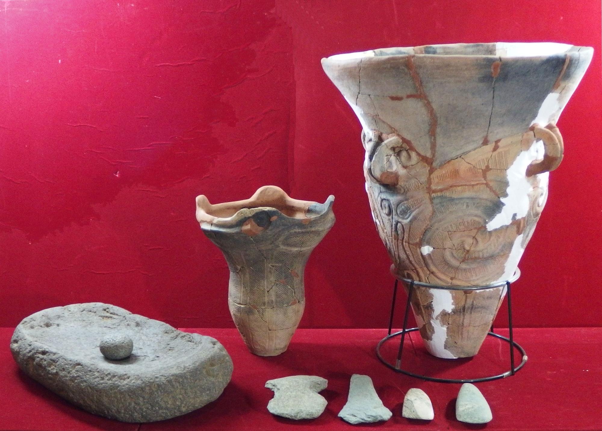 縄文時代の土器や石器が並べられており、左側には石皿とすり石が、中央には小型の土器が、右側には装飾的な大きな深鉢土器が金属製の台に置かれている写真