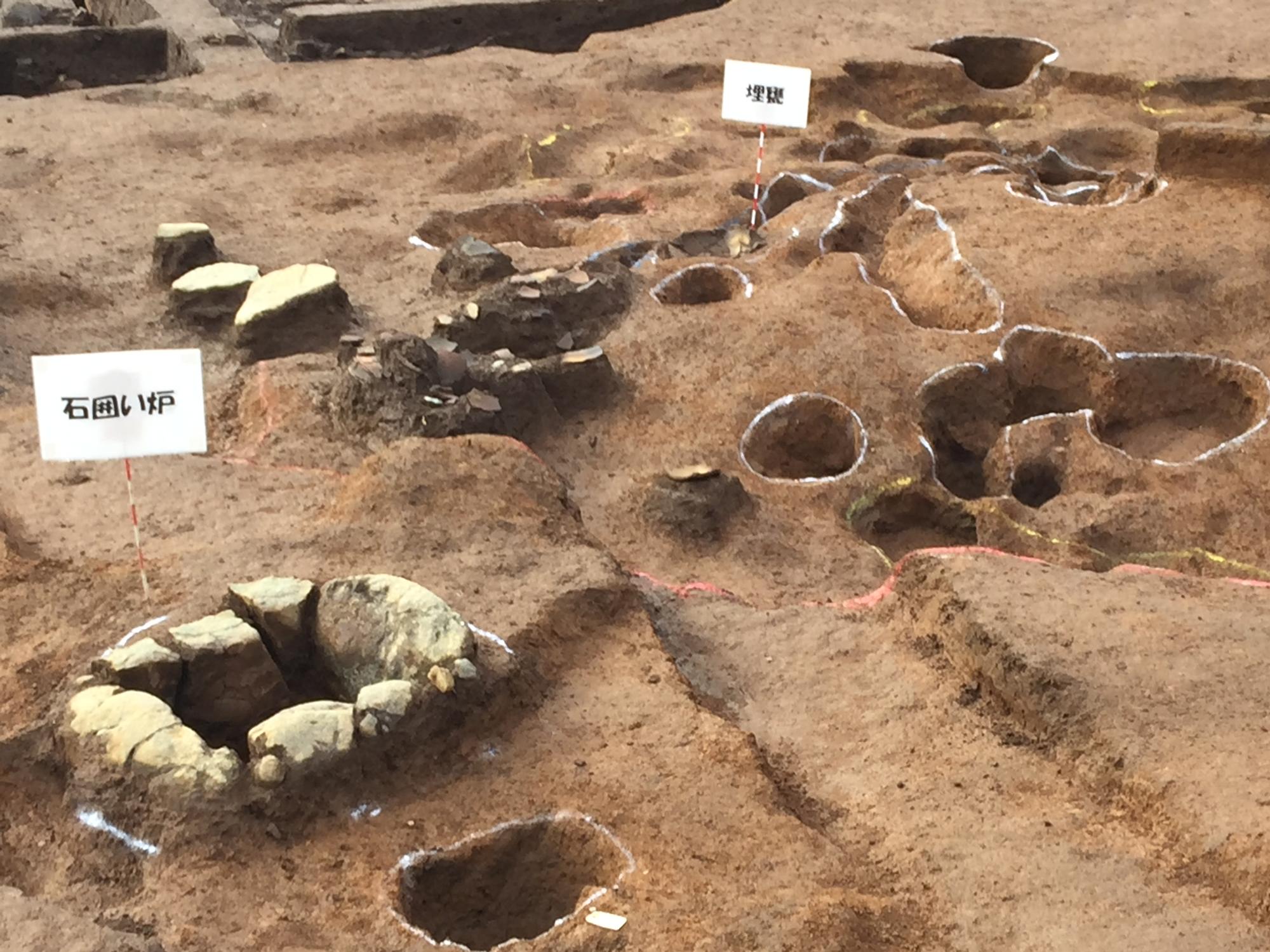 地面に複数の穴が開いた発掘現場で、周りを石で囲まれた穴の横と、写真の中央付近に文字が書かれた標識が立てられている様子の写真