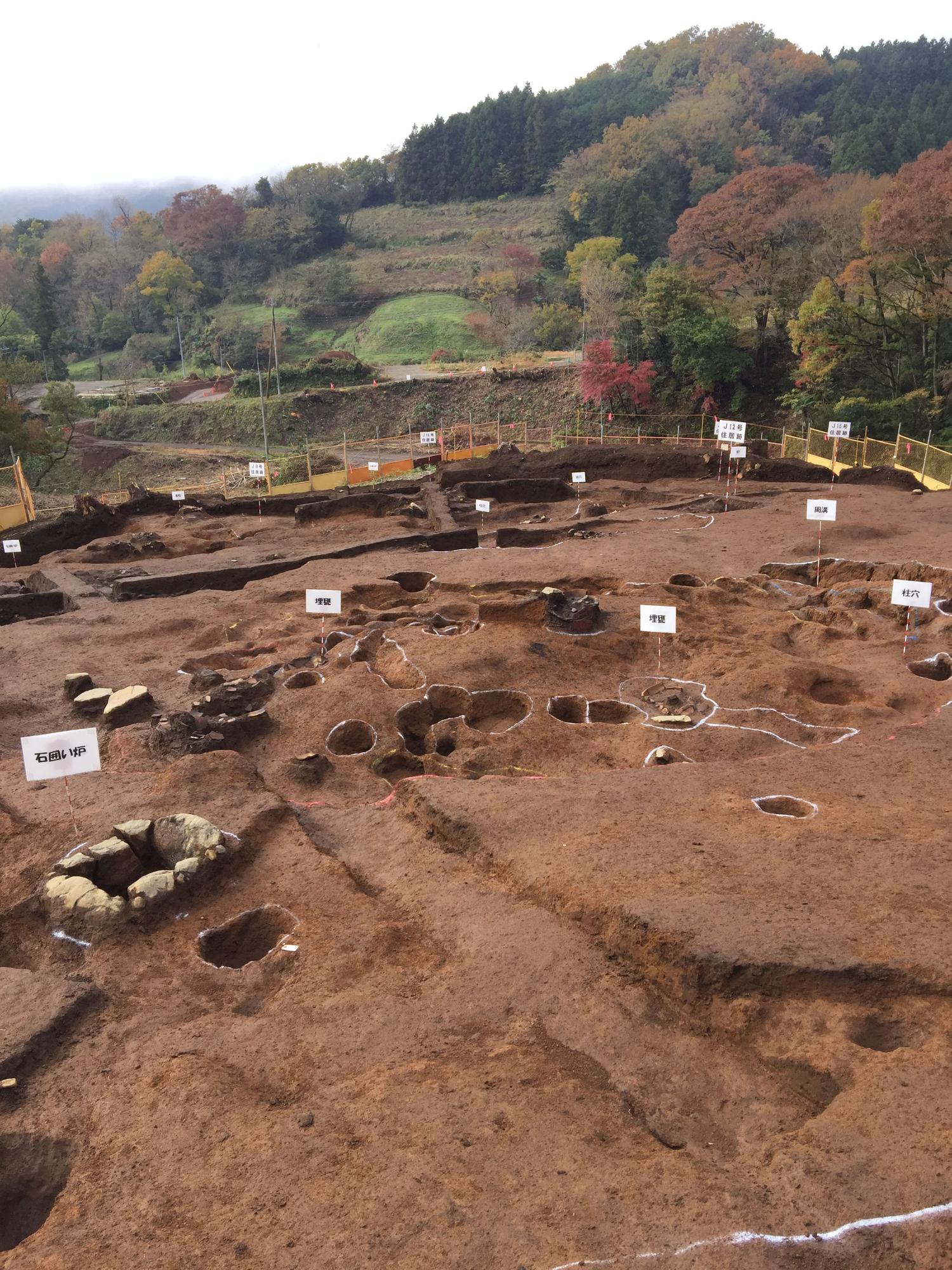 黄色いフェンスで囲まれた中に、たくさんの穴や石のようなものがあり、文字が書かれた白い小さな標識がたくさん立てられている、山のふもとの発掘現場の全景写真