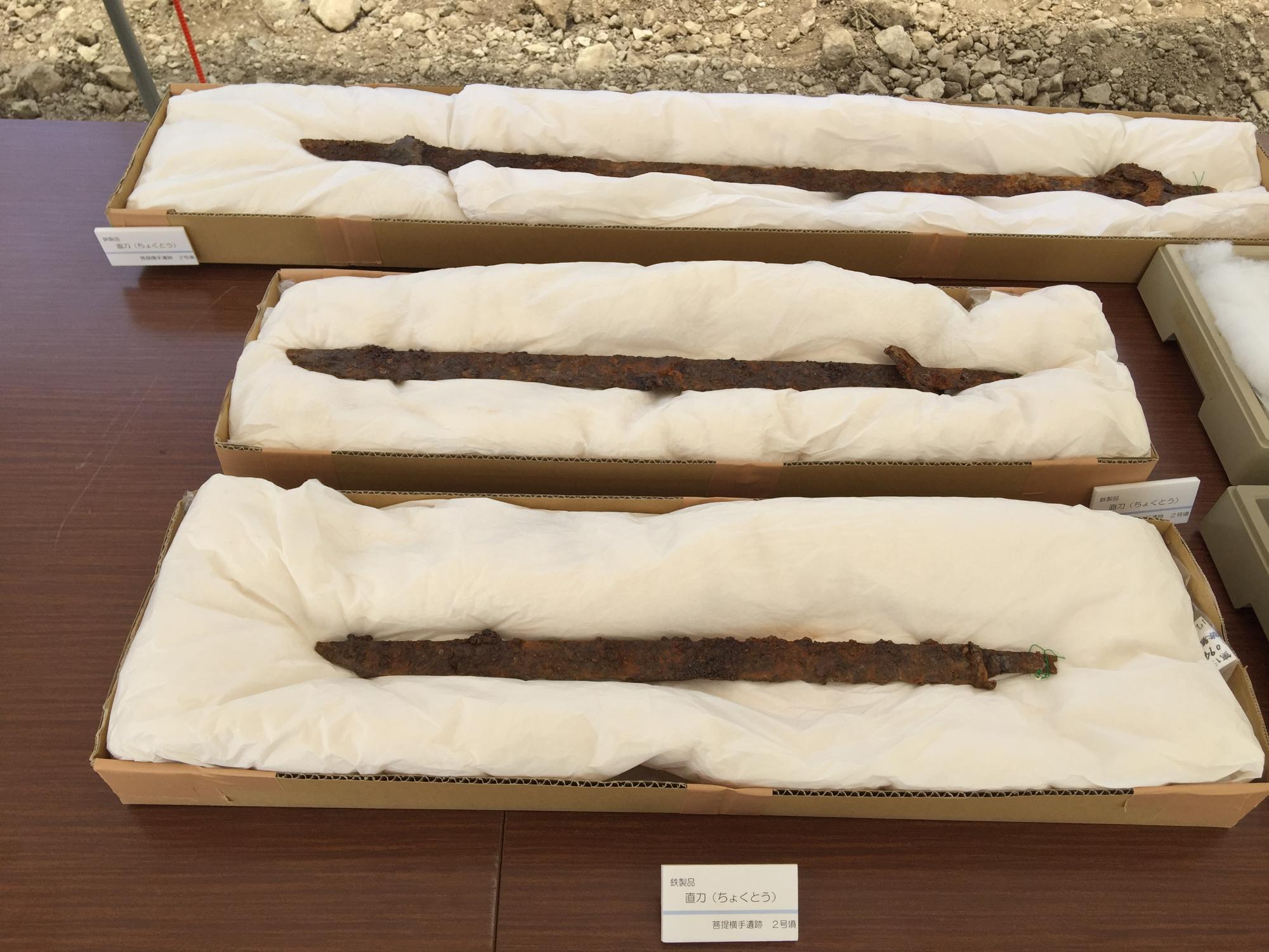 腐食し錆びついた3振りの直刀が、白い緩衝材を敷いた茶色の段ボール箱にそれぞれ納められ、木製のテーブルの上に一列に並べられて展示されている写真