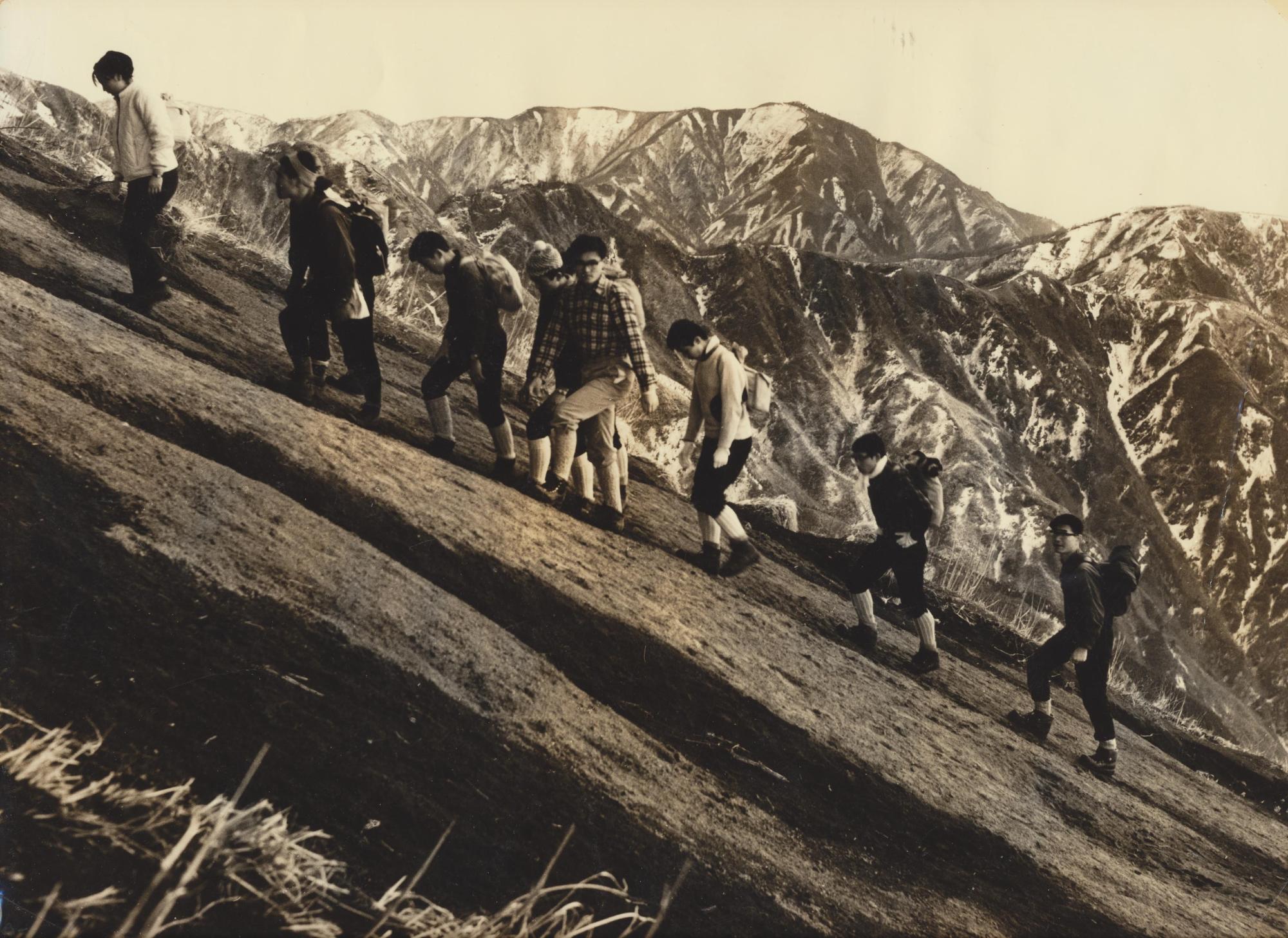 背後に山脈を臨む急な山肌を複数の登山者たちが登っている様子のモノクロ写真