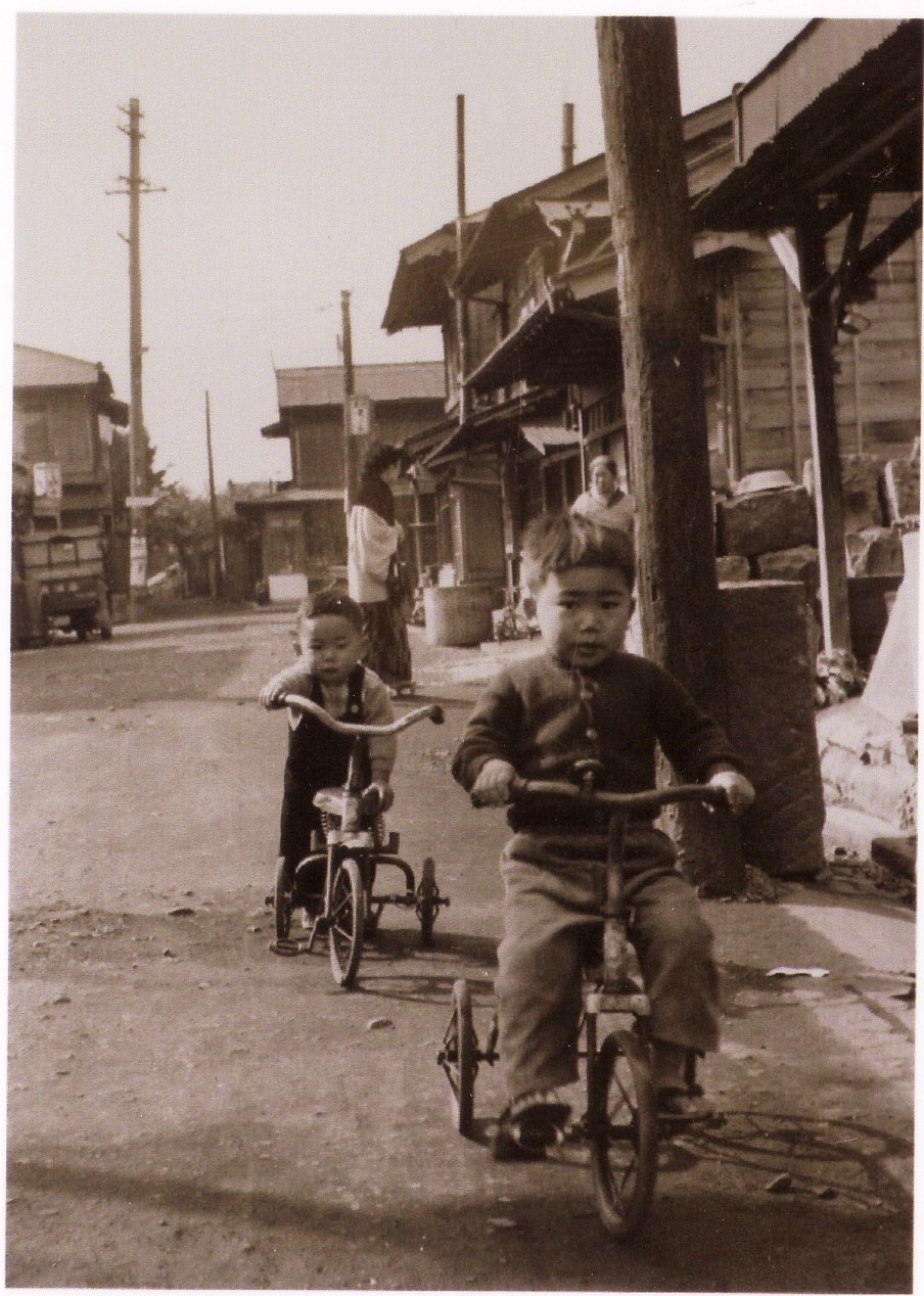 両側に木造の家が立ち並んだ通りを男の子2人がそれぞれ三輪車に乗って走っている様子のモノクロ写真