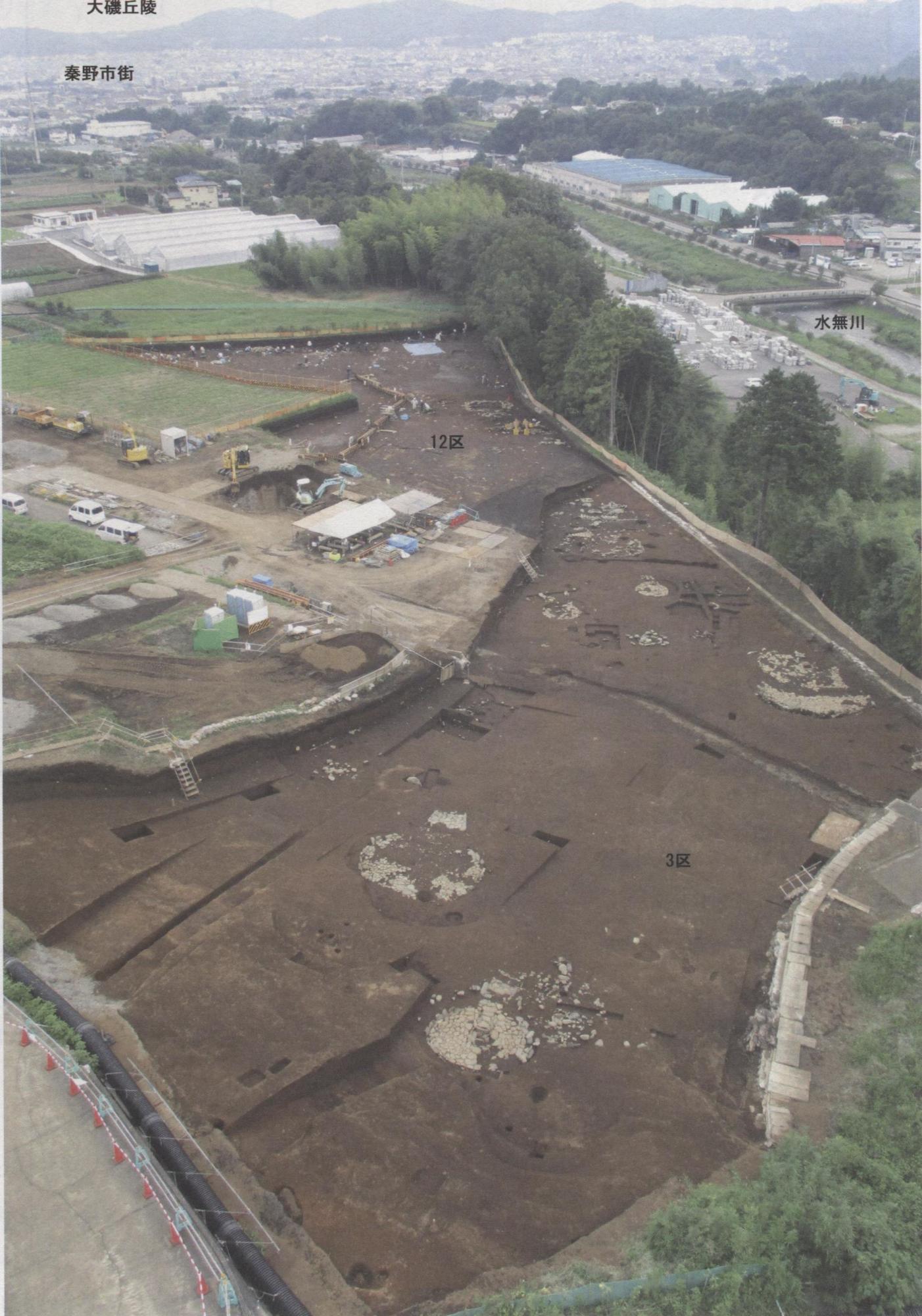 発掘調査中の大規模な遺跡を俯瞰で撮影した写真で、地面には円形の石組みや掘り出された複数の穴が見え、遠景には市街地や山が写り込んでいる稲荷木遺跡の写真