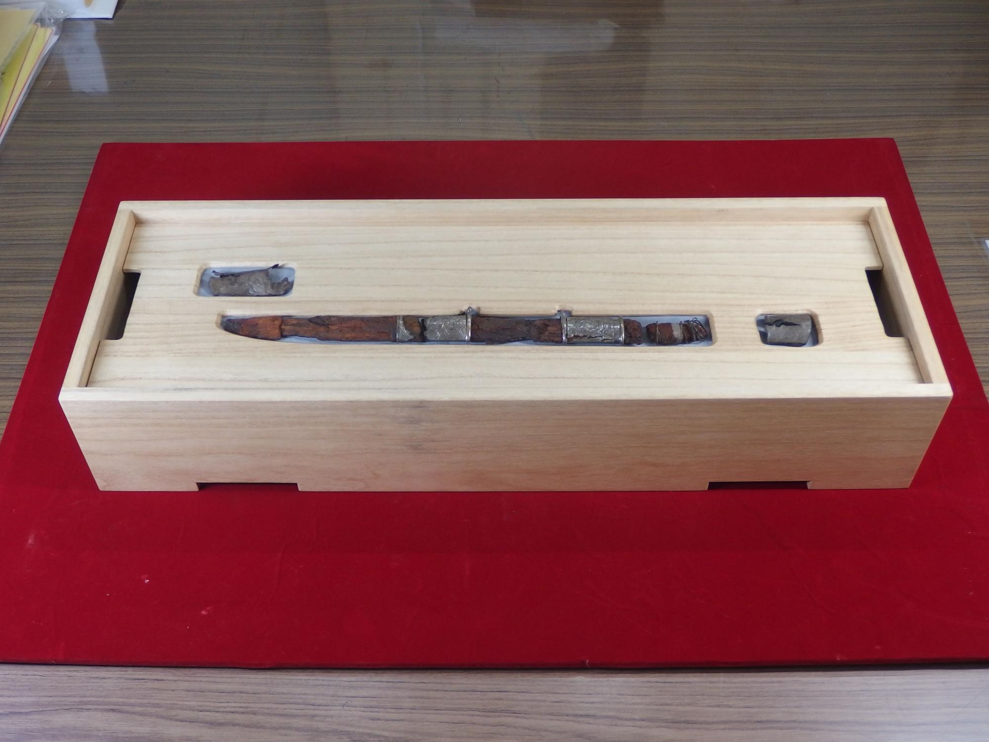 長方形の木箱の中に、古くて茶色い鞘と柄に銀色の装飾が施された刀剣のようなものが収められており、他の小さな断片も一緒に展示されている写真