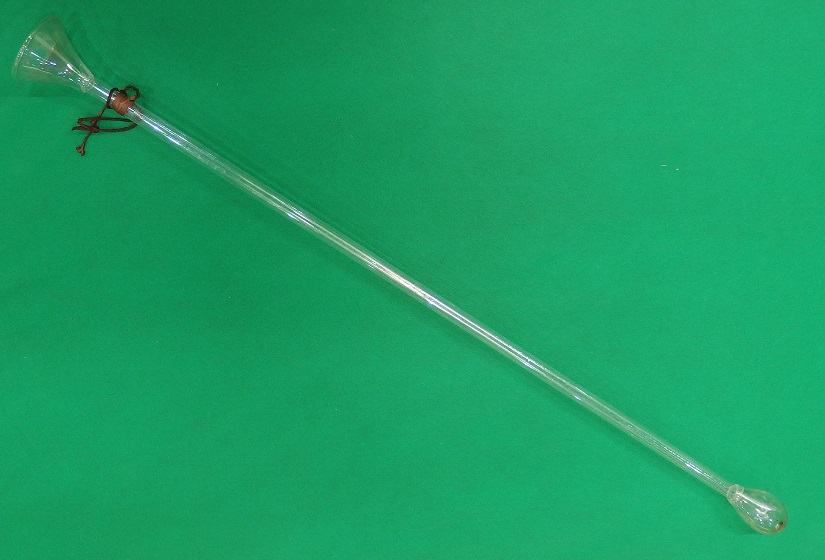 ラッパのような形のガラス部分と細長い管状のガラス部分が組み合わされた、全体が透明なガラスでできた珍しい形状のハエ取り器が緑色の布の上に置かれた写真