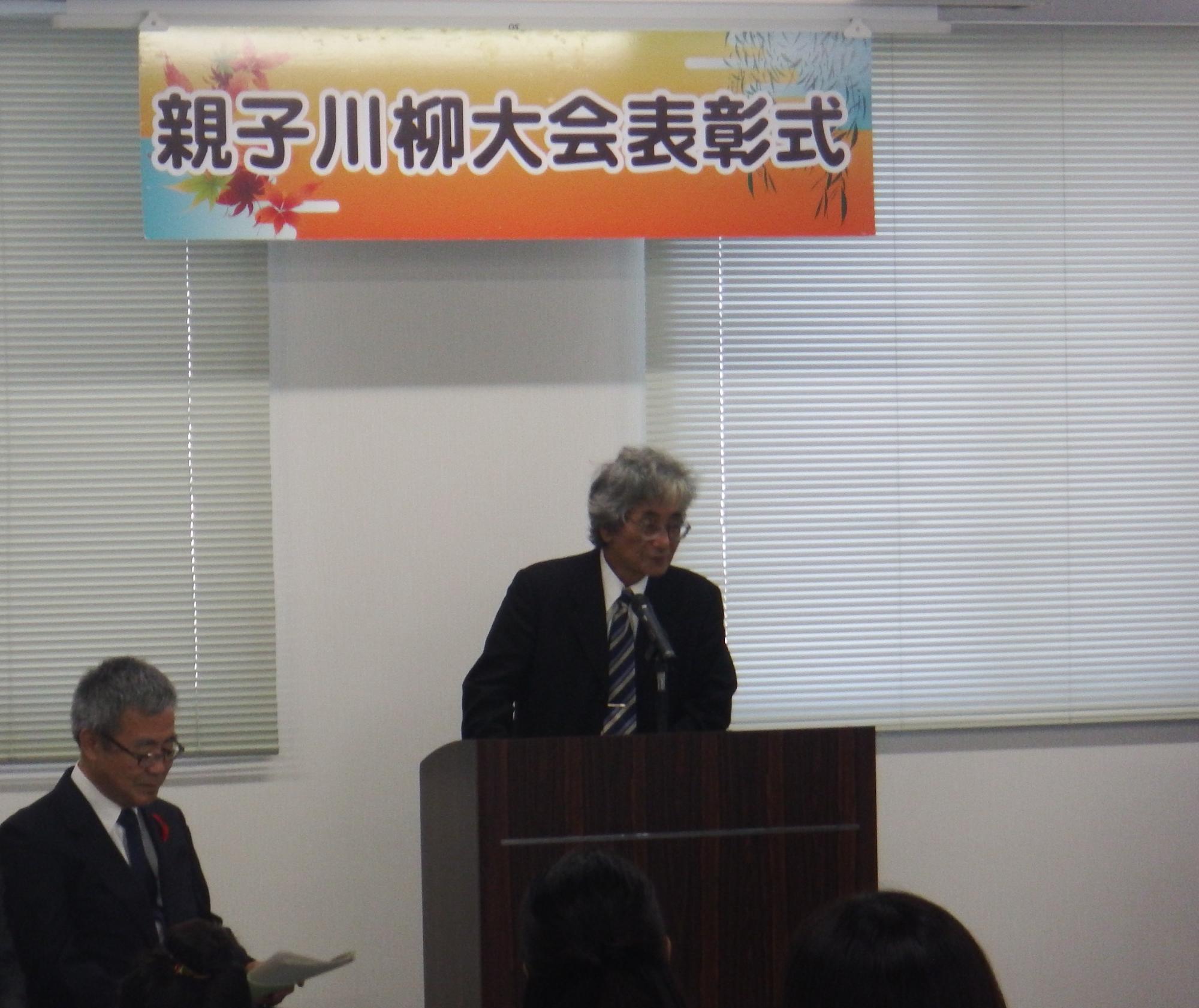 「親子川柳大会表彰式」と書かれた横断幕の下で、鍛冶実行委員長兼審査委員長が演台に立ち話をしている写真