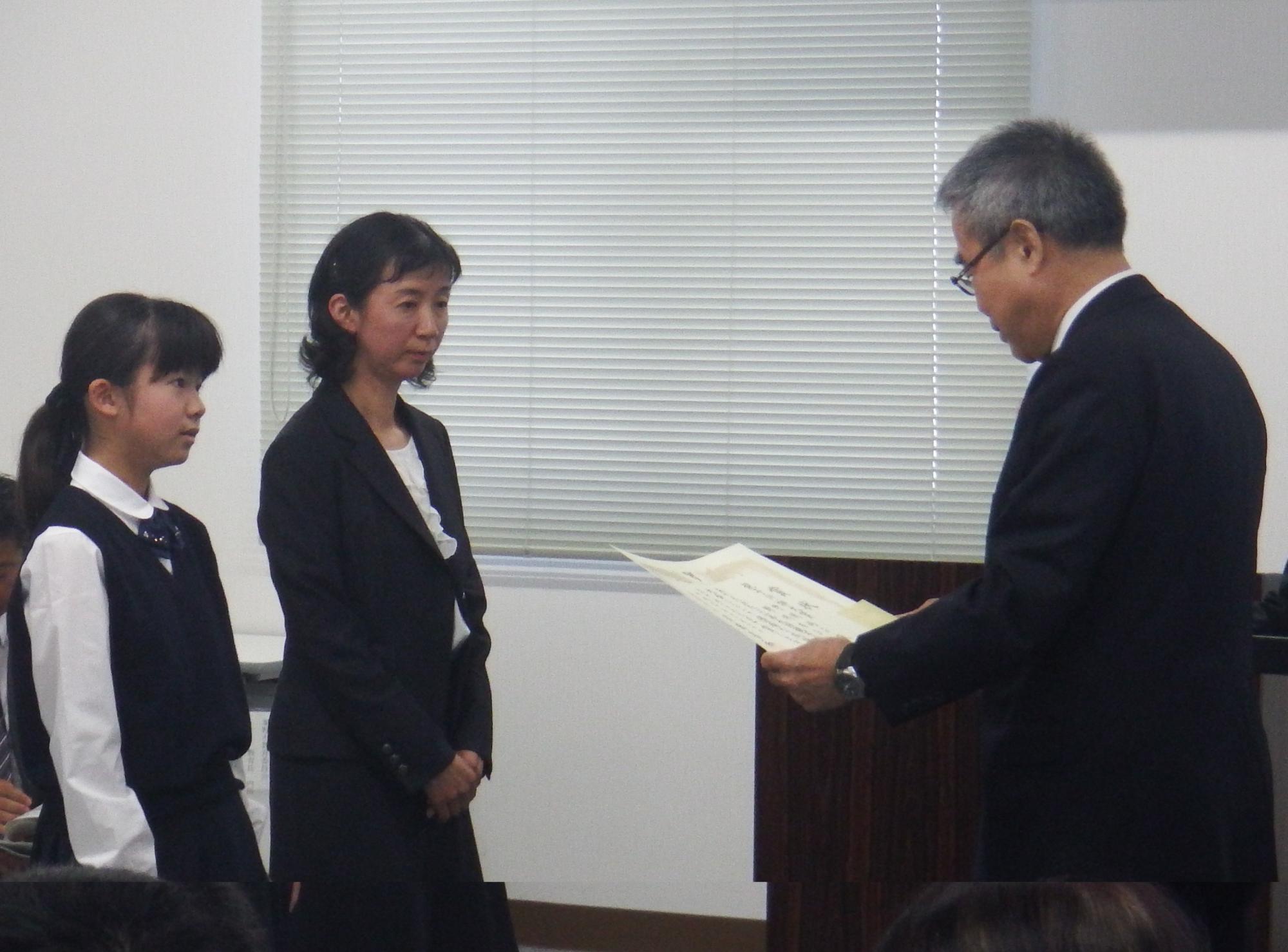 スーツ姿の男性が、制服を着た女児とその隣の女性に向かって表彰状を授与している様子を捉えた写真
