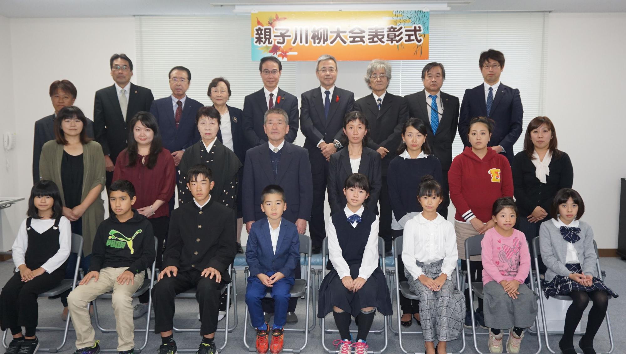 「親子川柳大会表彰式」の横断幕の下、受賞した児童とその家族、そして大会関係者や主催者と思われるスーツ姿の大人たちが、3列に整列している集合写真