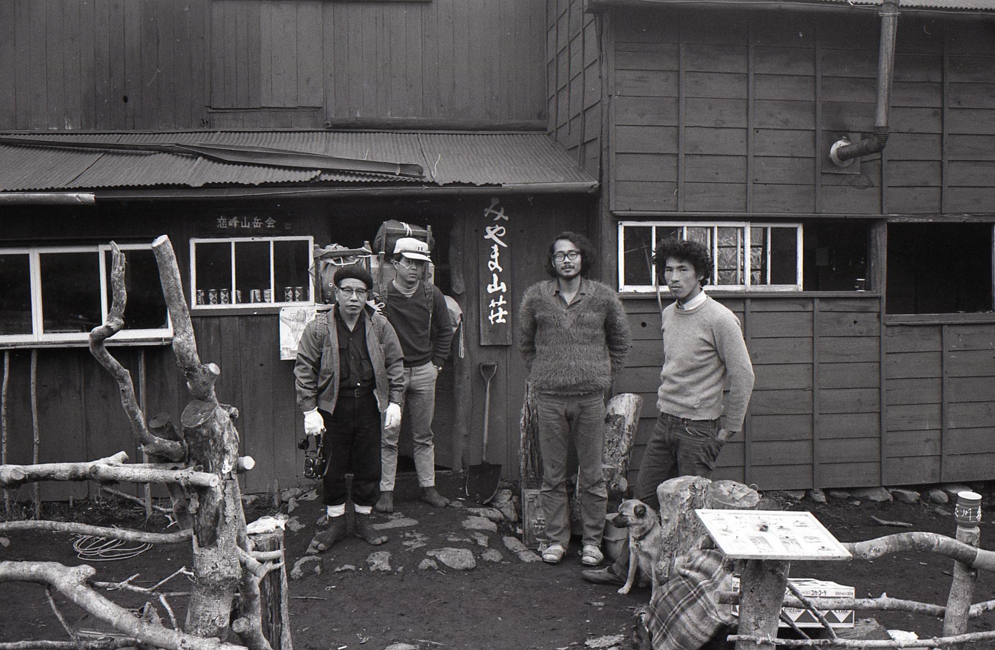 古い木造みやま山荘の前で、4人の男性が立ち、右側には一匹の犬がおり、その全員がカメラに向かって立っている様子を捉えた白黒写真